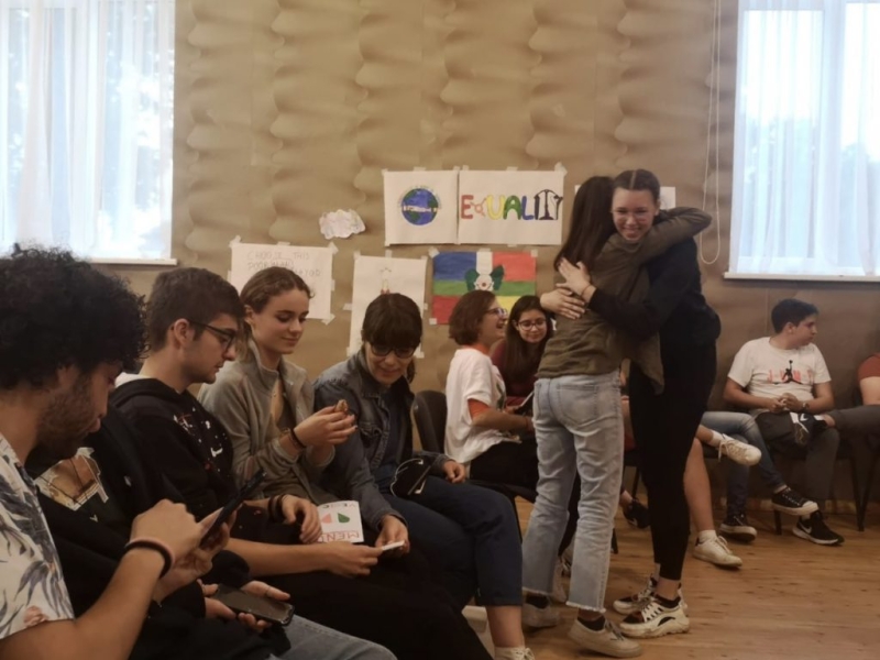 jaunieši sēž telpā uz krēsliem, divas meitenes apskaujas